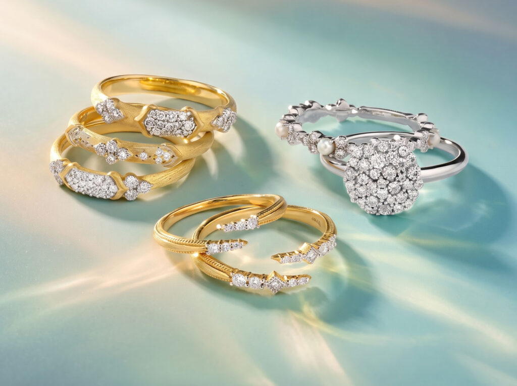 Stacked textured yellow gold bands with pavé diamonds and a white gold diamond cluster ring on a teal surface