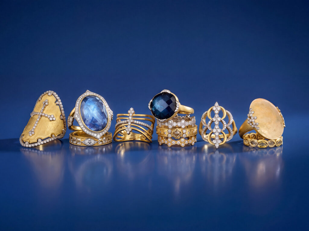 Ornate yellow gold statement rings set with diamonds, blue sapphires, labradorite, and citrine reflected on blue glass